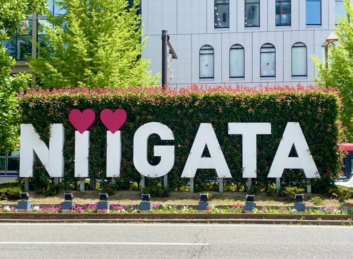 屋外に設置された「NIIGATA」デザイン看板。2つのハートが文字の I の代わりになっており、背景には緑の植栽と建物が見える。