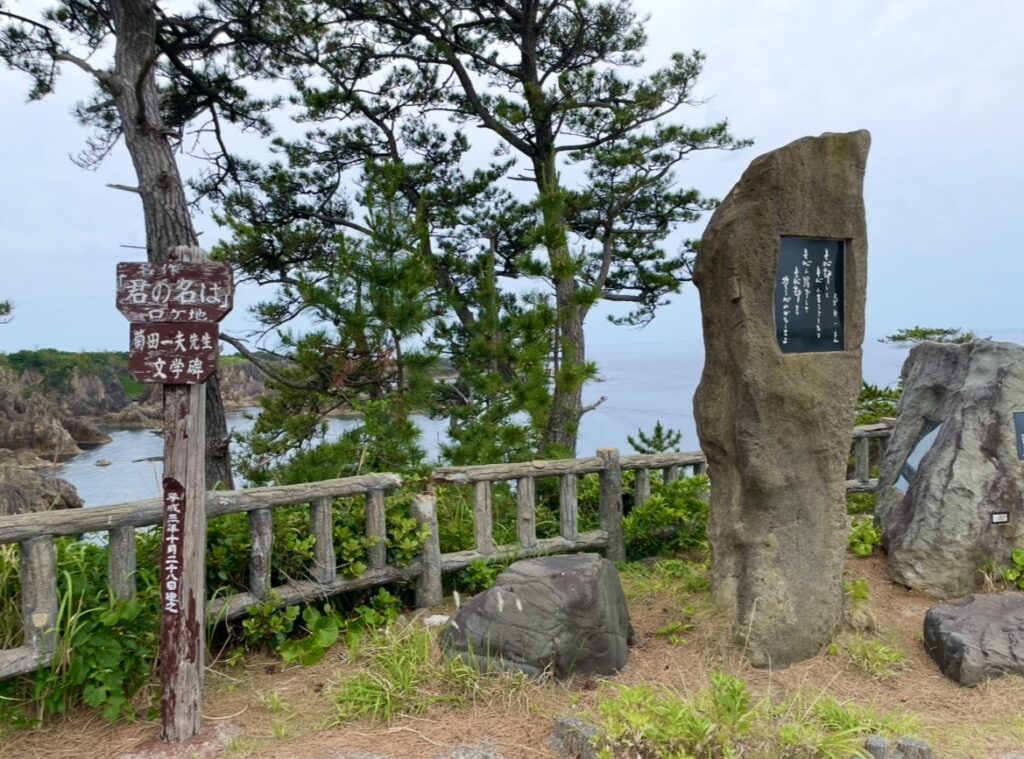 尖閣湾の展望スポットに立つ文学碑と案内板の写真。海岸の絶景を背景に、映画ロケ地としての記念碑が設置されている様子。