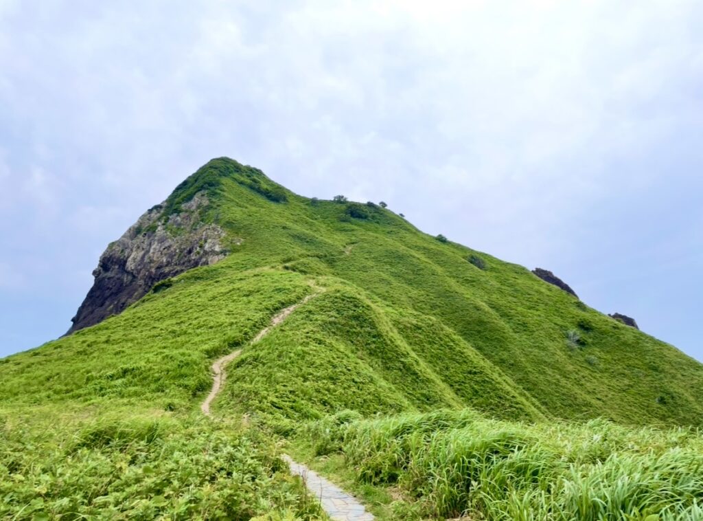 大野亀の山頂へ続く細い遊歩道が、緑に覆われた斜面をまっすぐ登っていく風景。草原と岩肌が広がる静かな山の写真。