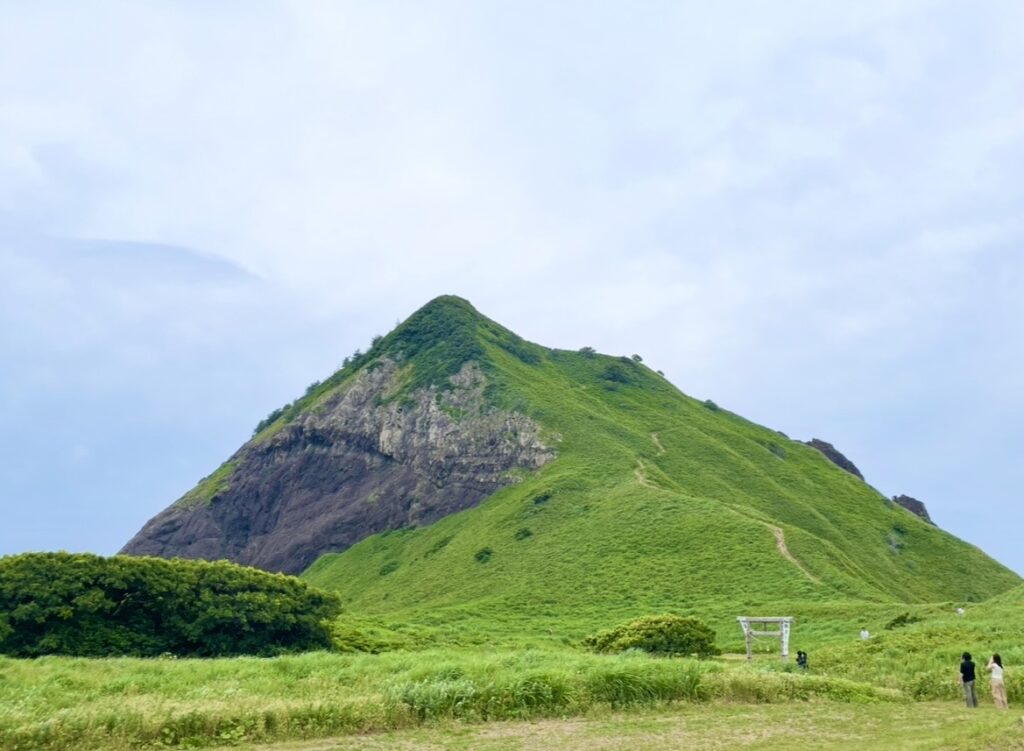 大野亀の巨大な一枚岩の山と、草原が広がる麓の風景。山肌の岩と緑が美しく対比し、登山道や鳥居が見える雄大な景観の写真。