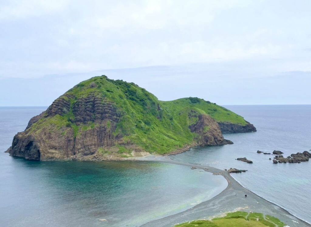 二ツ亀の島と本土を結ぶ砂州が海上に現れ、エメラルド色の浅瀬と深い青の海が美しく対比する風景。緑に覆われた岩山が海に浮かぶように佇む写真。