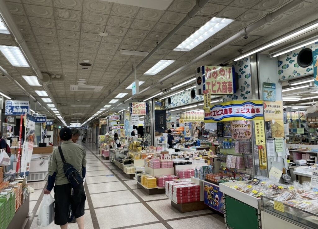 両津港のターミナル内に並ぶ土産店の売り場。棚いっぱいに商品が並び、買い物客が通路を歩くにぎやかな雰囲気の写真。