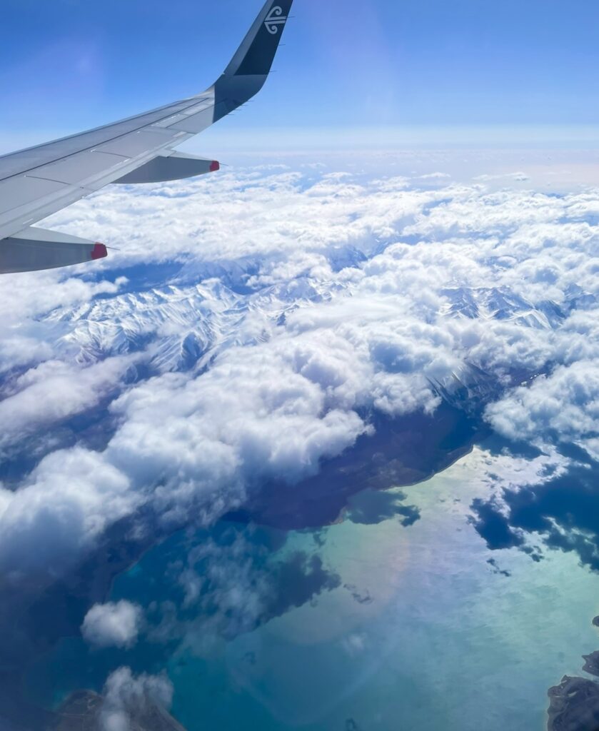 クイーンズタウン行きのニュージーランド航空便から撮影した大自然の空撮。雪をかぶった山々と雲海、青く輝く湖が広がる絶景を飛行機の翼越しに捉えた写真。