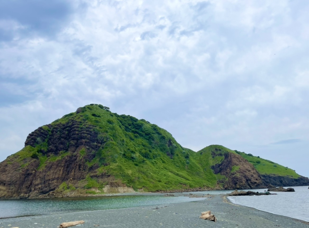 二ツ龜的岩山向海面延伸，前方是一片深色沙灘，覆滿綠意的陡坡直通海岸，寧靜海面與雲層天空相互映襯。