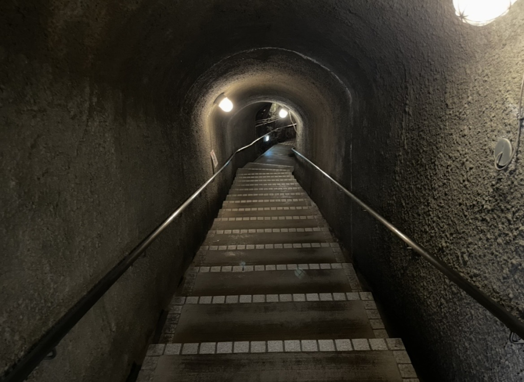 Interior of the Sado Gold Mine tunnel. A dim underground passage with continuing stairways, rocky walls, and spaced lighting that conveys the atmosphere of the Sōdayu Tunnel.