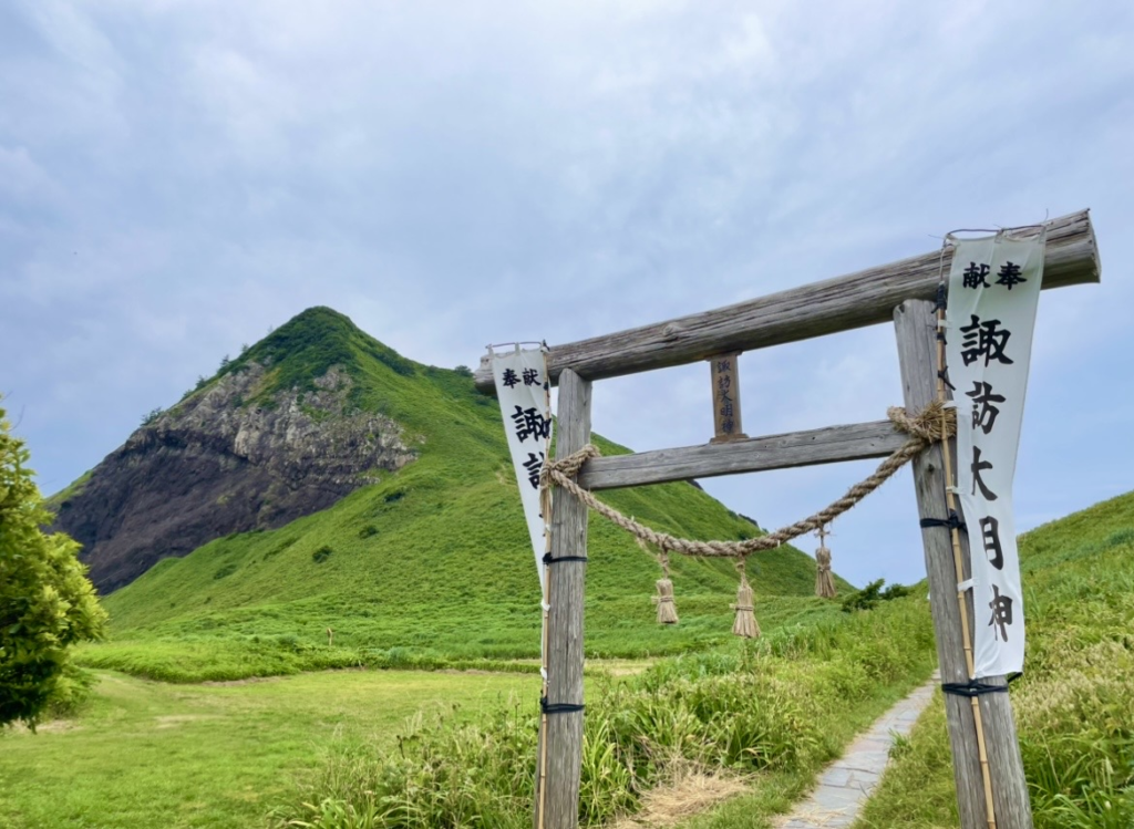 Torii gate and banners standing at the foot of Ōnogame, set against the mountain’s lush green slopes. The path leading upward conveys a solemn atmosphere where nature and local faith come together.