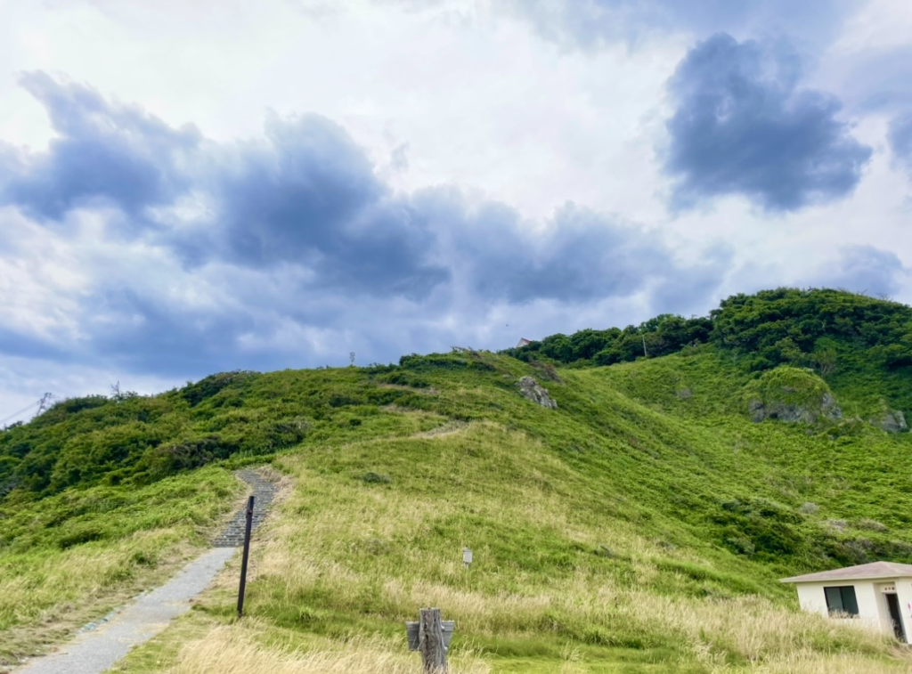 Narrow path descending the slope toward Futatsugame, surrounded by grass-covered hills. Heavy clouds fill the sky, and a small white lodge-like building stands quietly at the base.