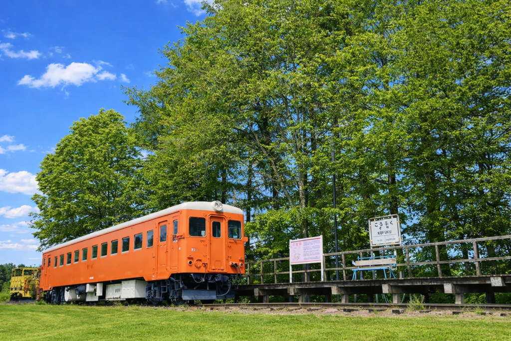 Under a clear blue sky, an orange diesel railcar sits on the platform of Kōfuku Station, surrounded by greenery.
The wooden platform and the station sign reading “Kōfuku / KOFUKU” stand side by side, creating a quiet, nostalgic scene where travelers can still feel the charm of this former station.