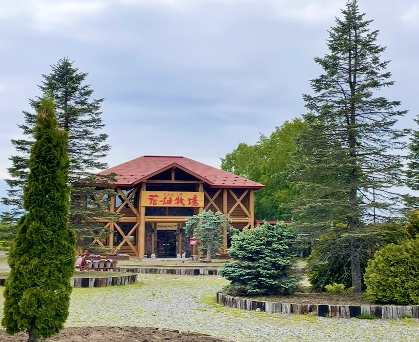 A wooden ranch building with a red roof, featuring a sign reading “Hanabatake Farm” above the entrance.
Trees and planted greenery surround the facility, set within a calm and pastoral countryside landscape.