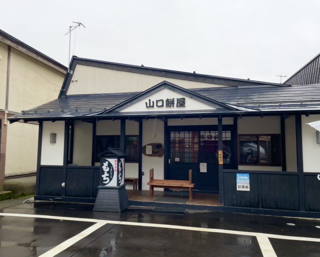 蔵王温泉にある山口餅屋の外観。青い屋根と木造の店構えが特徴で、入口前にはベンチと「もち」と書かれた看板が並ぶ。雨で濡れた石畳が温泉街らしい雰囲気を引き立てる老舗の和菓子店。