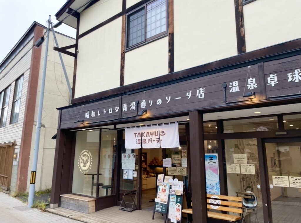 蔵王温泉にあるTAKAYUパーラーの外観。木造のレトロな店構えに「温泉パーラー」の暖簾が掛かり、昭和レトロ風の看板が並ぶ。ガラス越しにカウンターやメニューが見える、高湯通りの人気カフェスポット。