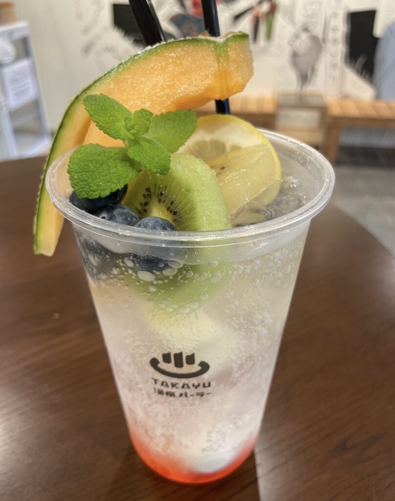 蔵王温泉のTAKAYUパーラーで提供されるTAKAYUフルーツソーダ。キウイ、レモン、メロン、ブルーベリー、ミントが入った彩り豊かな炭酸ドリンクで、透明カップにTAKAYUのロゴが入る人気メニュー。