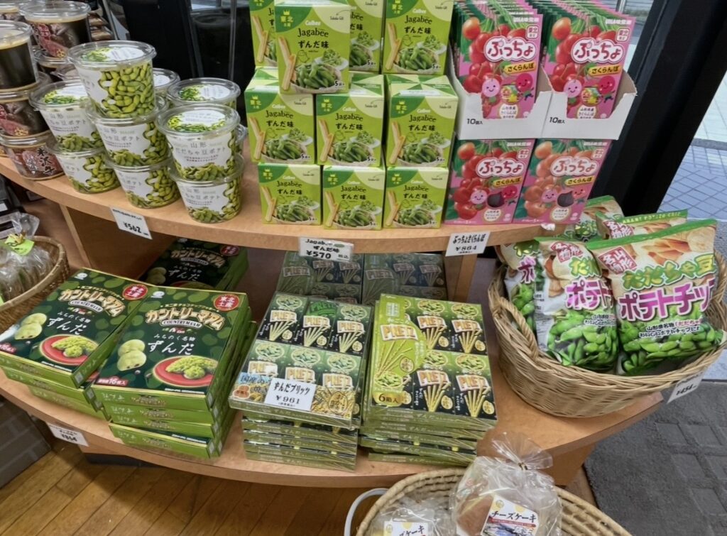 土産店の棚に並ぶずんだ味のお菓子類。ずんだプリッツ、ずんだカントリーマアム、ずんだ味ジャガビーなど緑色のパッケージ商品が並び、山形名物のずんだを使った人気土産が一面に陳列されている。