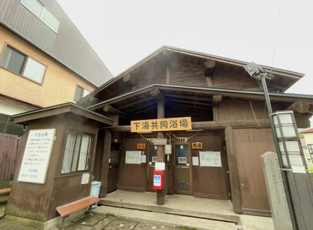 蔵王温泉の下湯共同浴場の外観。木造の趣ある建物に男女別の入口が並び、赤いポストや案内板、ベンチが置かれた昔ながらの共同浴場。温泉街の歴史を感じるレトロな雰囲気が漂う。