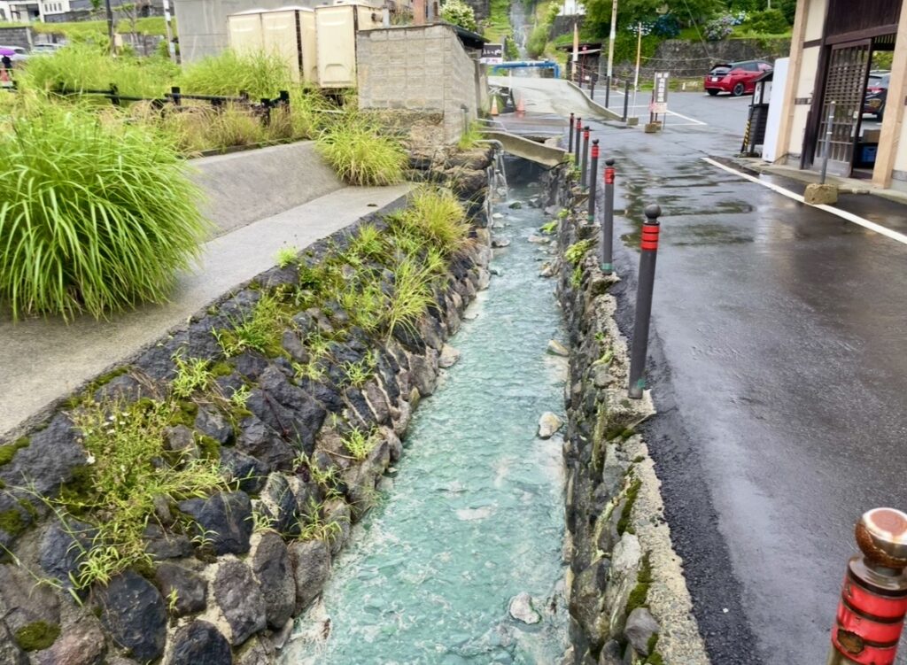 蔵王温泉街を流れる温泉の水路。青みがかった温泉水が石造りの細い水路を流れ、周囲には草木が生える。道路沿いに赤いポールが並び、温泉地特有の景観が広がる。