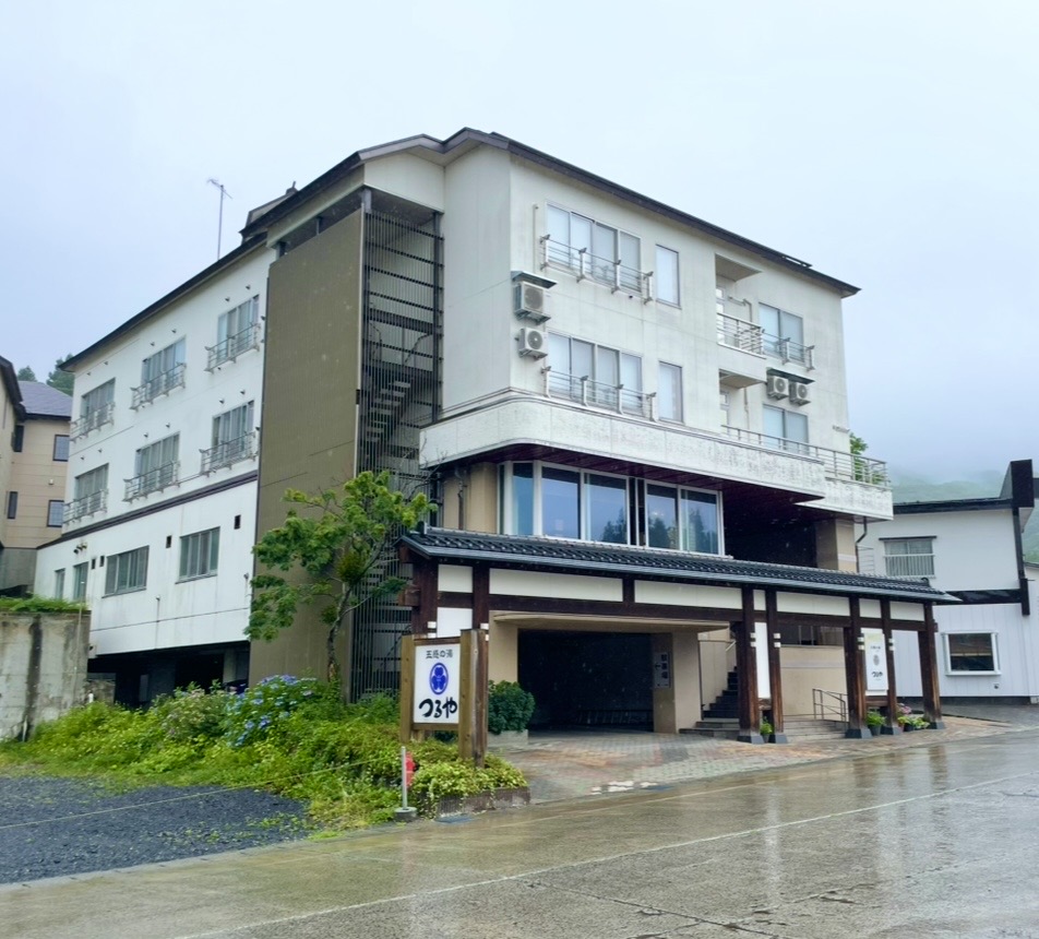 蔵王温泉の旅館「五感の湯 つるや」の外観。白壁の建物に木製の玄関が設けられ、雨に濡れた石畳と周囲の緑が温泉街らしい落ち着いた雰囲気を演出する老舗宿。