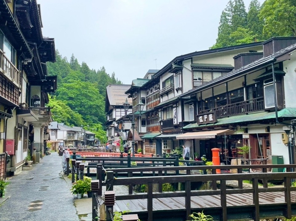 A small river flowing through the center of Ginzan Onsen, lined with wooden ryokan. People walk across the black‑railed bridge and along the stone‑paved path, while the deep greenery in the background harmonizes with the scene, highlighting the Taisho‑era romantic charm of the hot spring town.