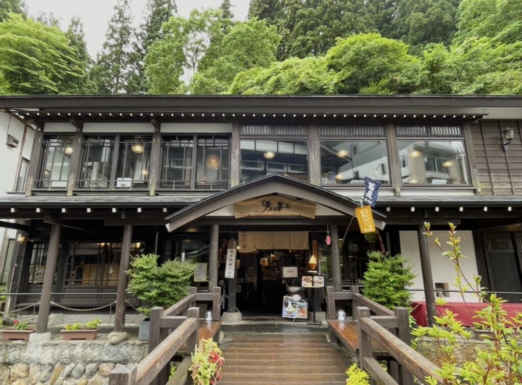 銀山温泉の名店「伊豆の華」の外観と入口の橋が調和する風景。緑に囲まれた静かな環境に、暖簾や看板が並ぶ和の趣ある施設が佇む