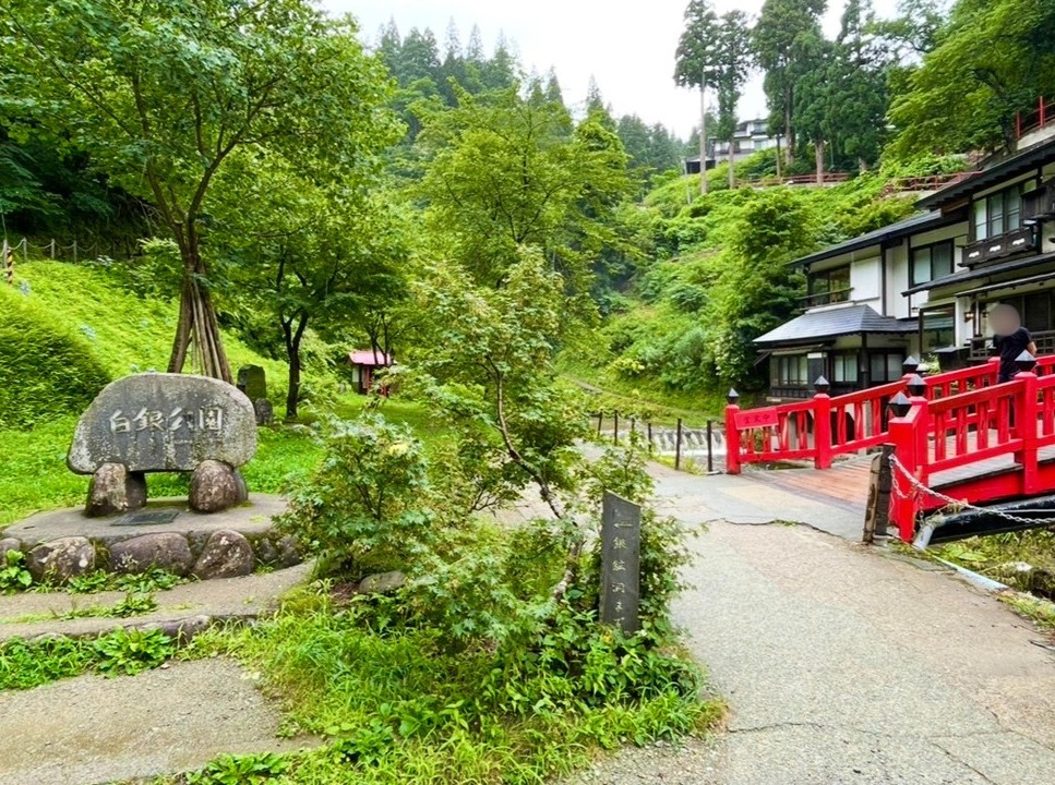 Stone monument and red bridge in Shirogane Park, surrounded by lush greenery. A quiet park area where traditional buildings and a small stream blend harmoniously with the natural setting.