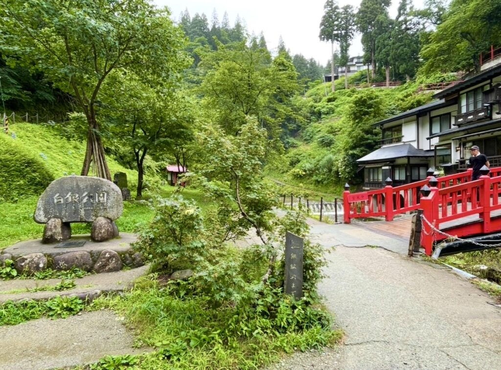 白銀公園の石碑と赤い橋が並ぶ風景。緑豊かな木々に囲まれ、伝統的な建物と小川が調和する静かな公園エリア。