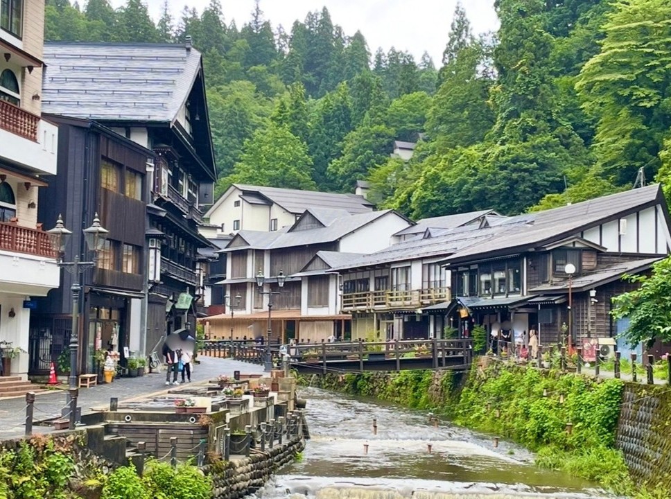 Wooden ryokan lined along the river in Ginzan Onsen, with tourists walking on the stone‑paved path. The historic buildings on both sides blend with the deep greenery in the background, creating a quiet and serene atmosphere typical of a traditional hot spring town.