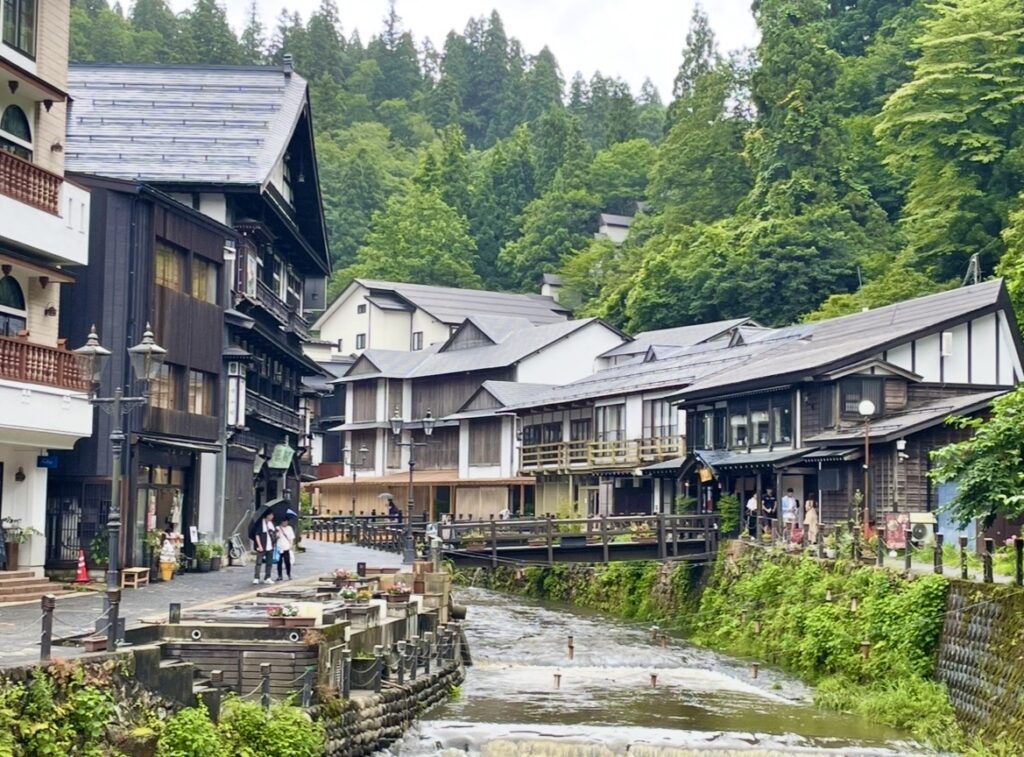 銀山温泉の川沿いに並ぶ木造旅館群。石畳の道を歩く観光客と、両側の歴史ある建物が調和し、背後の深い緑が温泉街らしい静かな雰囲気をつくる風景。