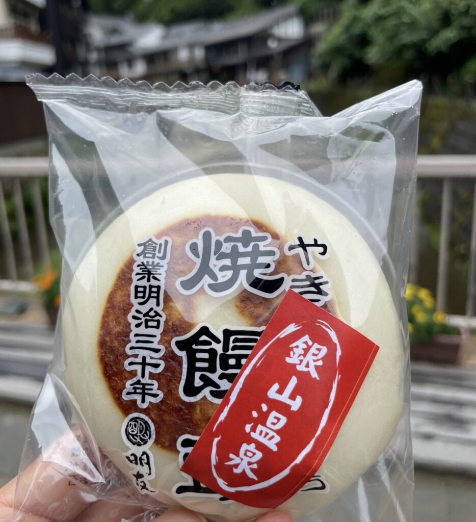 銀山温泉・西塚菓子舗の焼饅頭を手にした様子。香ばしく焼き上げられた饅頭の包装と、背景に広がる温泉街の風景が名物菓子の魅力を引き立てる。