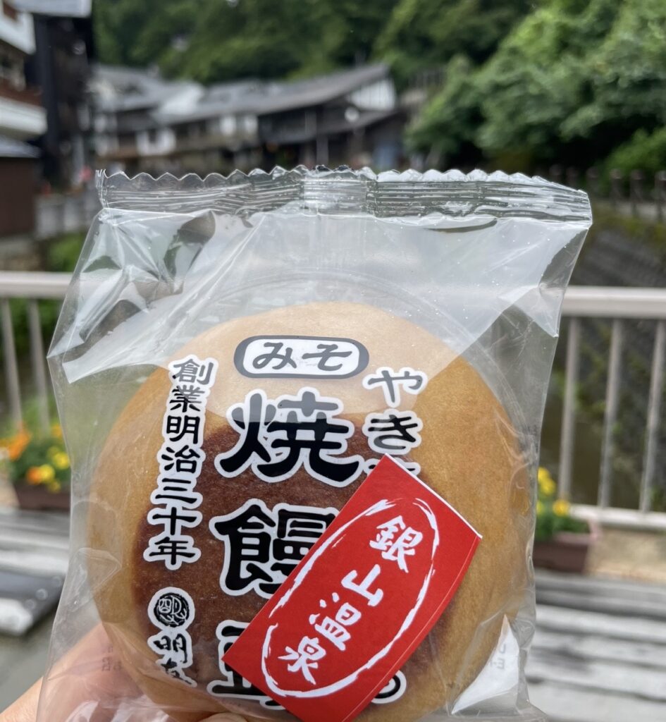 銀山温泉・西塚菓子舗の名物みそ焼饅頭を手にした様子。包装に刻まれた老舗の歴史と、背景に広がる温泉街の風景が名物菓子の魅力を引き立てる。
