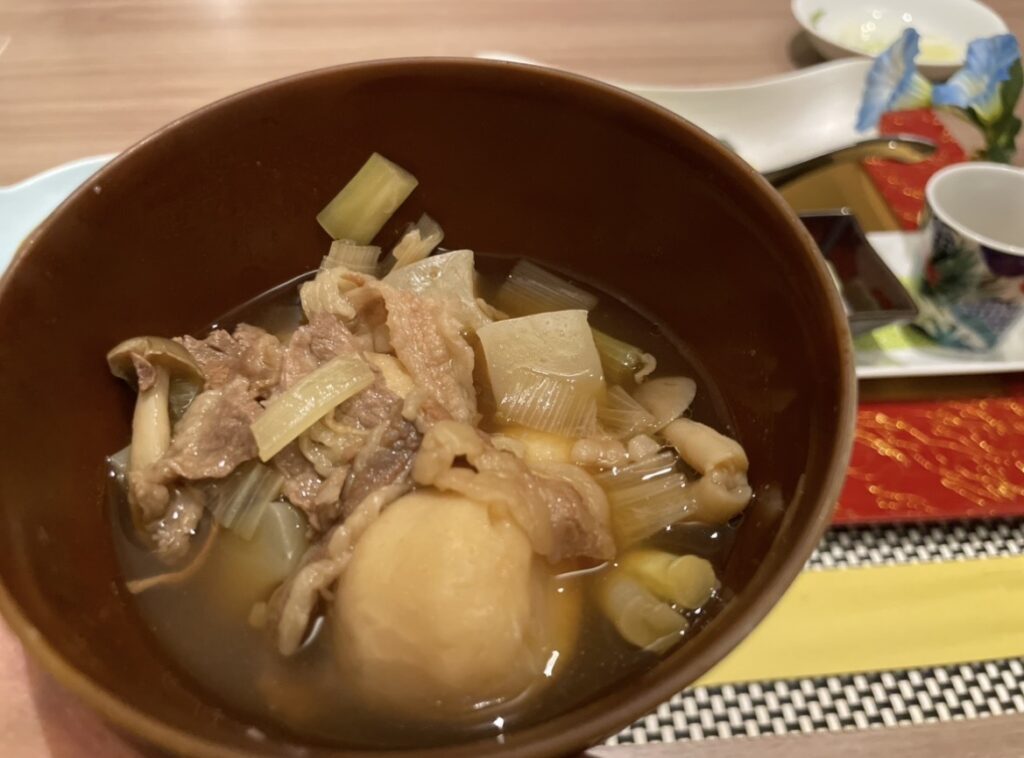 山形の郷土料理・芋煮の一杯。牛肉、里芋、長ねぎ、きのこが出汁の効いたつゆに煮込まれ、温かみのある器で提供される伝統の味わい。