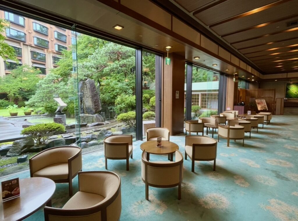 ほほえみの宿 滝の湯の中庭を望むソファー席。大きな窓越しに日本庭園の緑が広がる、くつろぎやすいスペースの写真。