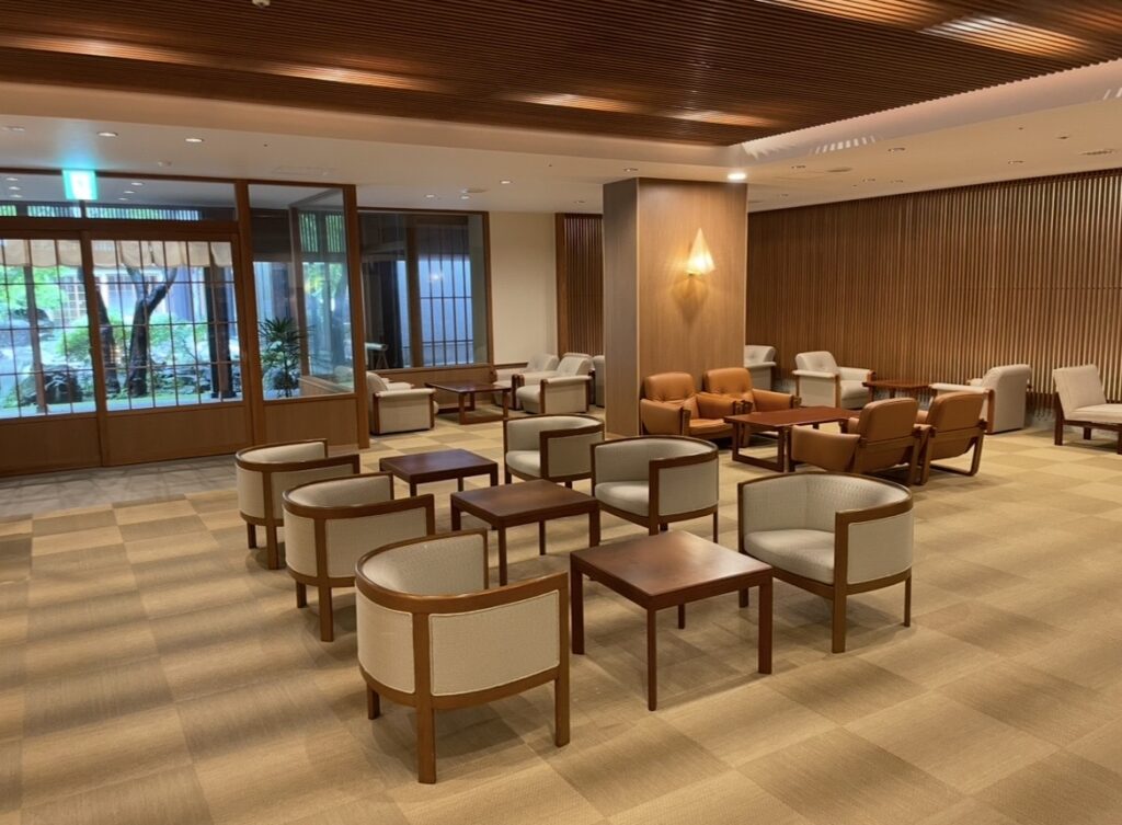 ほほえみの宿 滝の湯の館内ラウンジ。木製テーブルとソファが整然と並び、大きな窓から緑が見える開放的なラウンジスペースの写真。