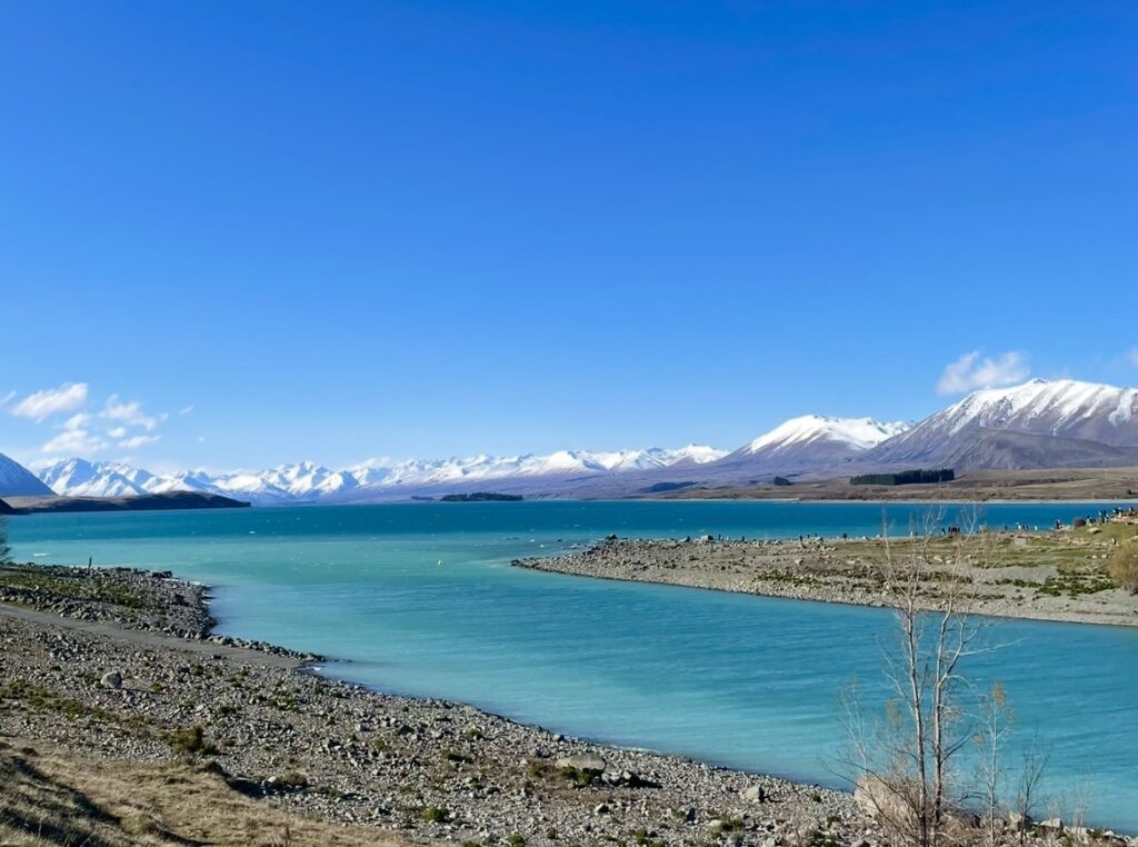 ミルキーブルーのテカポ湖と雪化粧した山々が広がる絶景。澄んだ青空の下、湖畔の岩場と透明な水面が輝くニュージーランド南島の自然風景