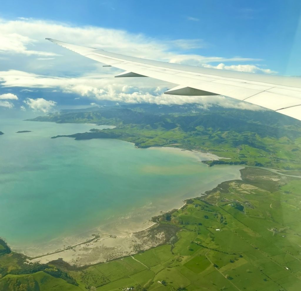 Aerial view of New Zealand’s coastline and lush green landscape seen over the airplane wing, with mountains covered in clouds and the blue ocean captured during an Air New Zealand flight.