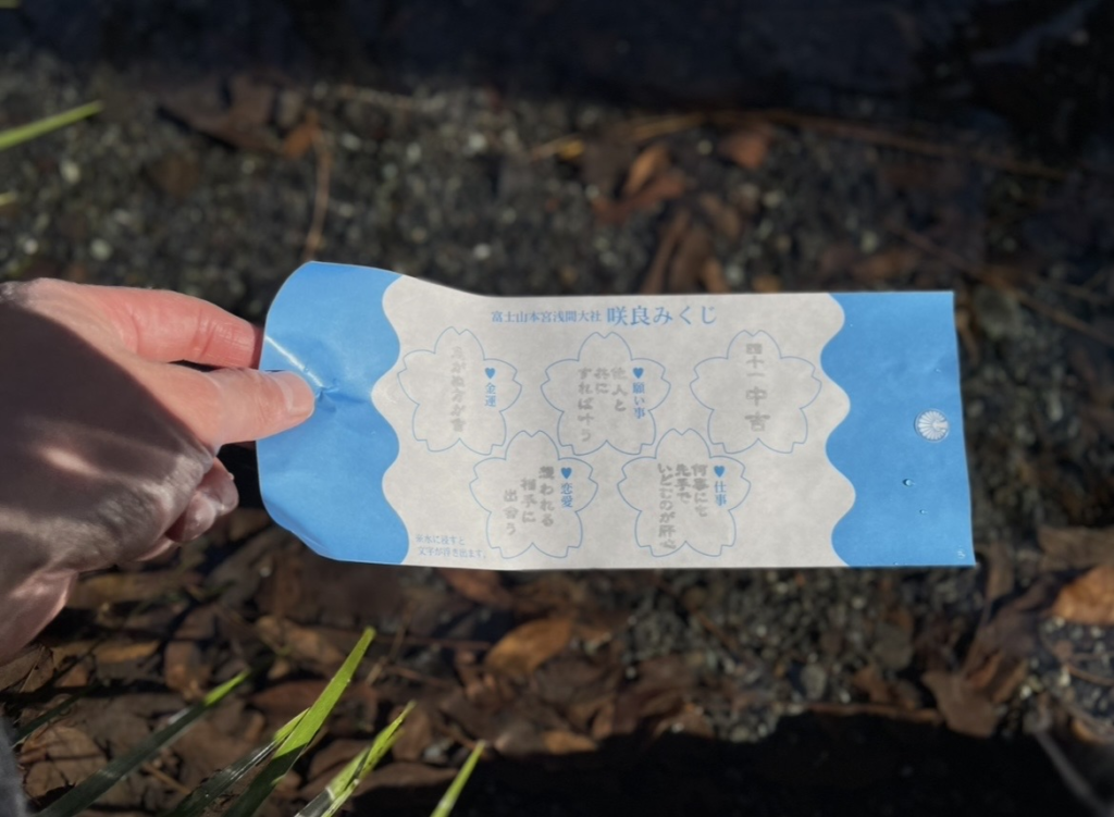 Sakura Omikuji from Fujisan Hongu Sengen Taisha, a flower‑shaped fortune slip that reveals its message when placed in sacred spring water, shown being held above the pond.