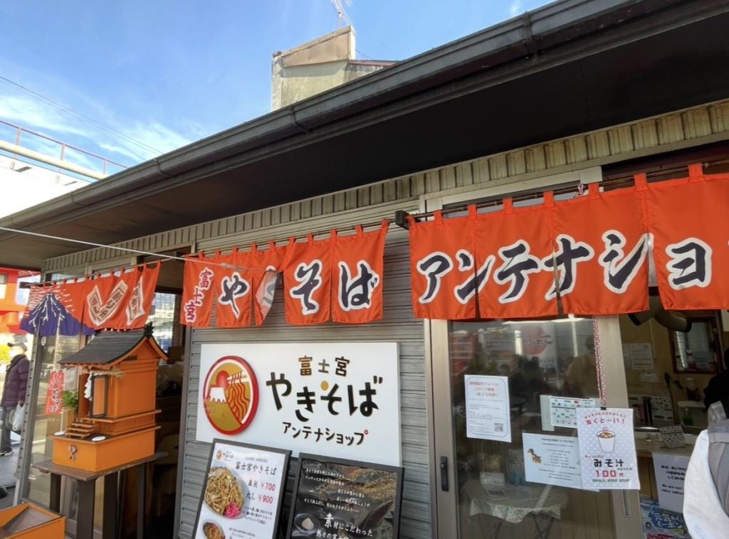 富士宮炒麵天線商店的店面,橘色布簾與招牌十分醒目,門口展示富士宮名物炒麵的菜單,並可見以富士山為意象的裝飾。