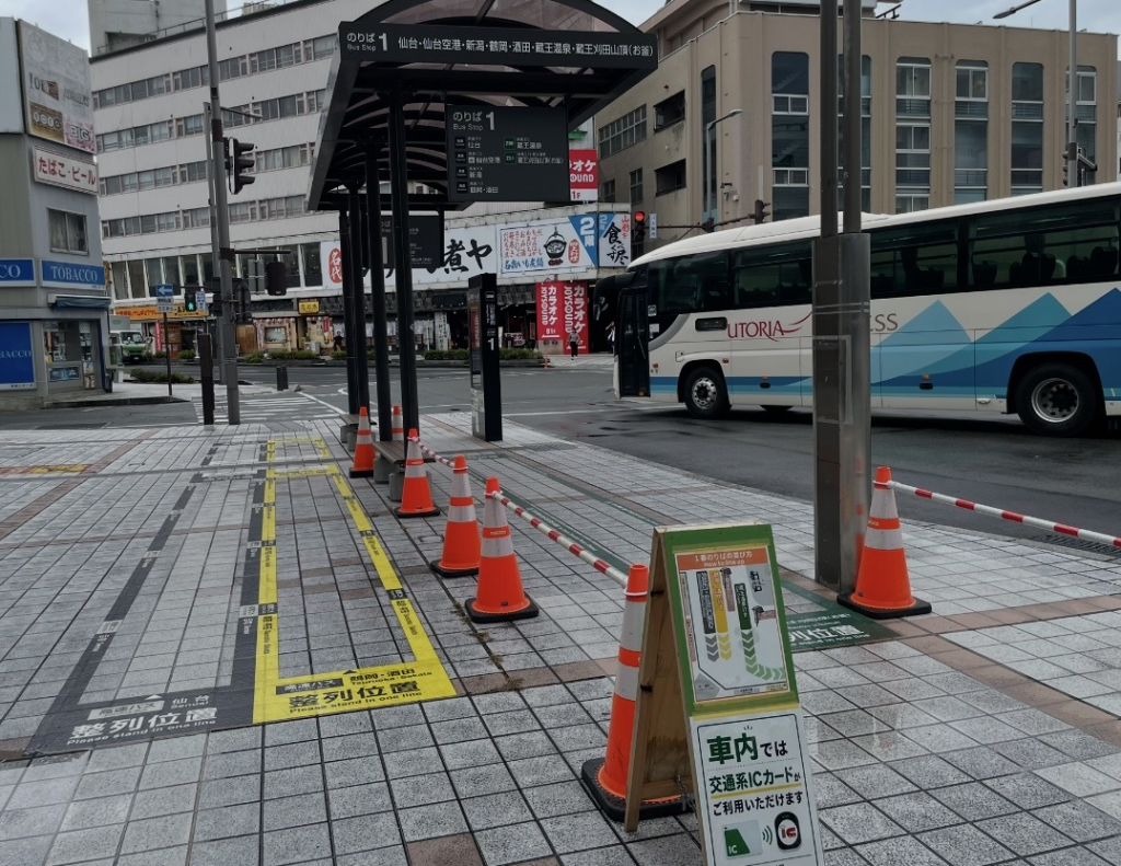 A view of the bus terminal in front of Yamagata Station, showing Bay 1 with its covered boarding area and a highway bus bound for Sendai and Sendai Airport. Clear signage indicates the boarding line and the availability of IC transit cards, with nearby shopping streets visible around this key transportation hub.