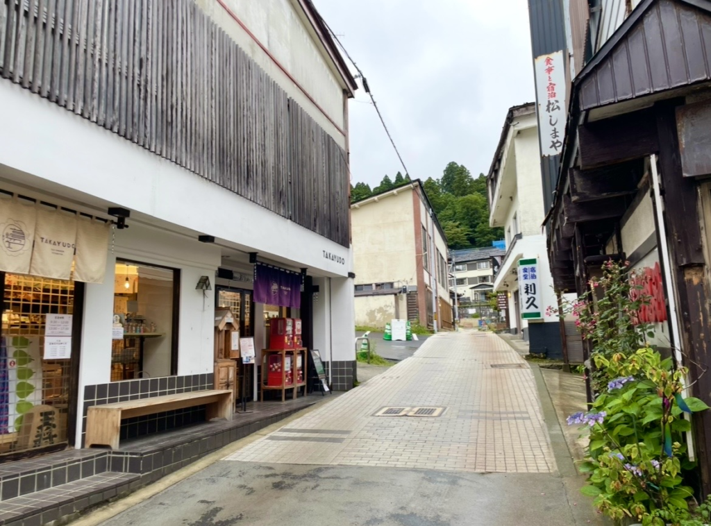 高湯通寧靜的街景。
細長的街道兩旁是傳統建築，店前的暖簾、木製長椅與和風招牌清晰可見。
石板路延伸至街道深處，周圍的綠意與花朵營造出溫泉街特有的悠閒氛圍。
