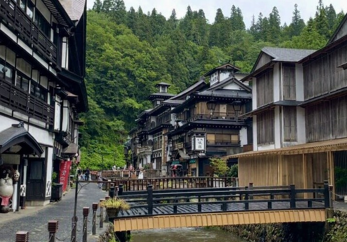 Retro streetscape of Ginzan Onsen, where a small river flows through the center of the hot spring town lined with wooden buildings. A black‑railed bridge and stone‑paved walkway extend along the water, harmonizing with the forested backdrop to create a Taisho‑era romantic atmosphere at this popular sightseeing spot.