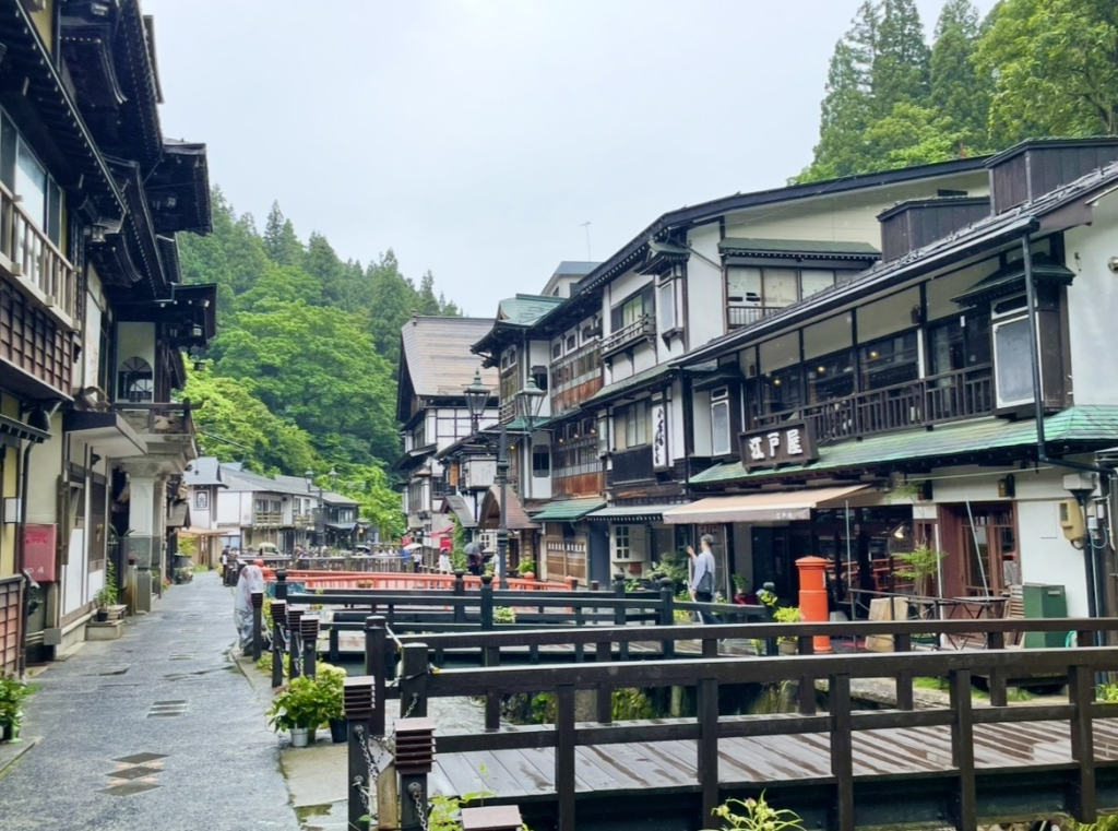 銀山溫泉中央流過的小河與兩側的木造旅館景致。
人們走在黑色欄杆的橋上與石板路間，背後濃密的綠意相互融合，呈現出大正浪漫氛圍的溫泉街魅力。