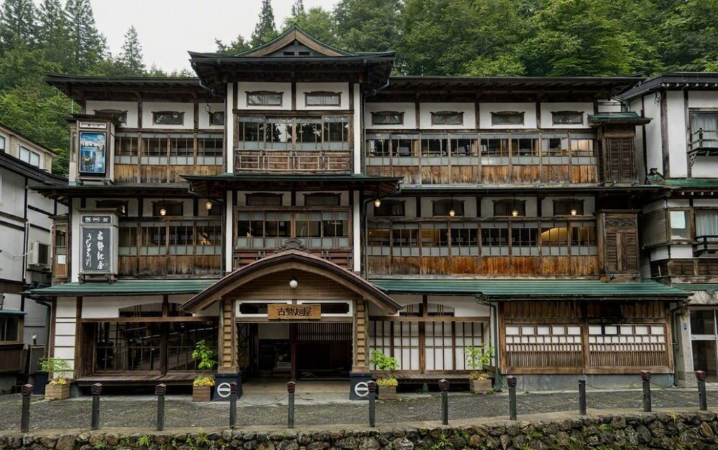 Historic wooden ryokan in Ginzan Onsen, featuring lattice windows and a stately roofline. Surrounded by greenery, the traditional architecture harmonizes with the stone‑paved street, creating a Taisho‑era romantic atmosphere throughout the hot spring town.