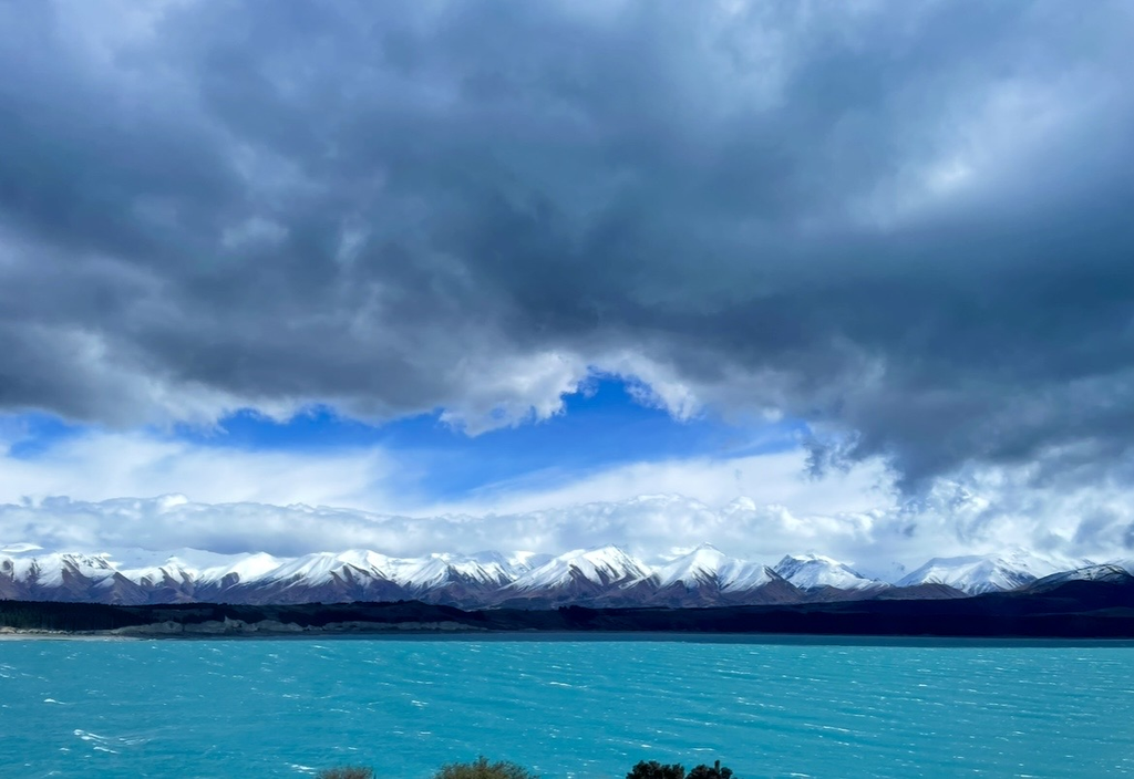 A dynamic landscape of milky‑blue Lake Tekapo, snow‑covered mountains, and dramatic clouds spreading across New Zealand’s South Island.