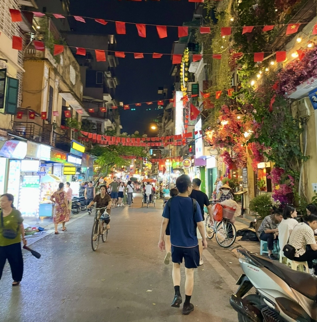 Lively night scene in Hanoi’s Old Quarter with rows of red flags and people and bicycles moving through the street
