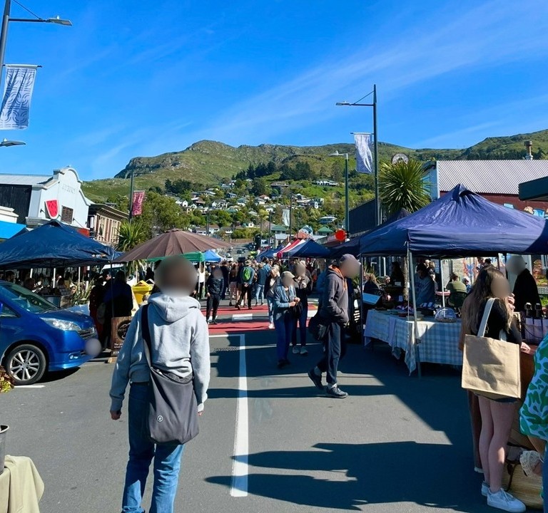 A lively scene from the farmers’ market in Lyttelton, Christchurch. Under the blue sky, local residents shop for produce and handmade goods at the bustling outdoor stalls.