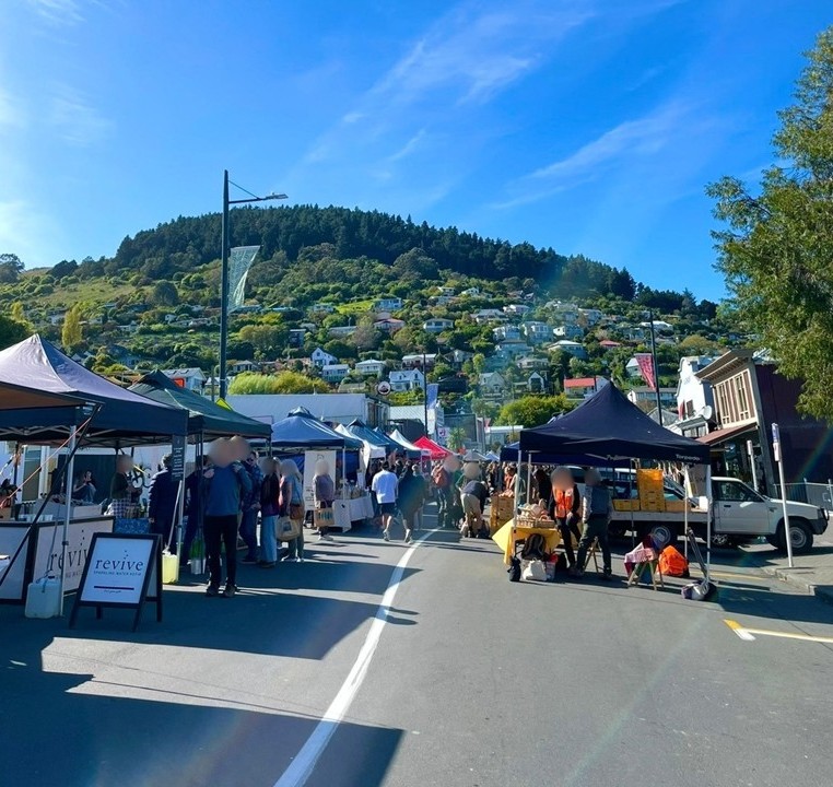People walking along a street lined with stalls at the farmers’ market in Lyttelton, Christchurch. A lively local atmosphere spreads under the blue sky with views of the surrounding hills.