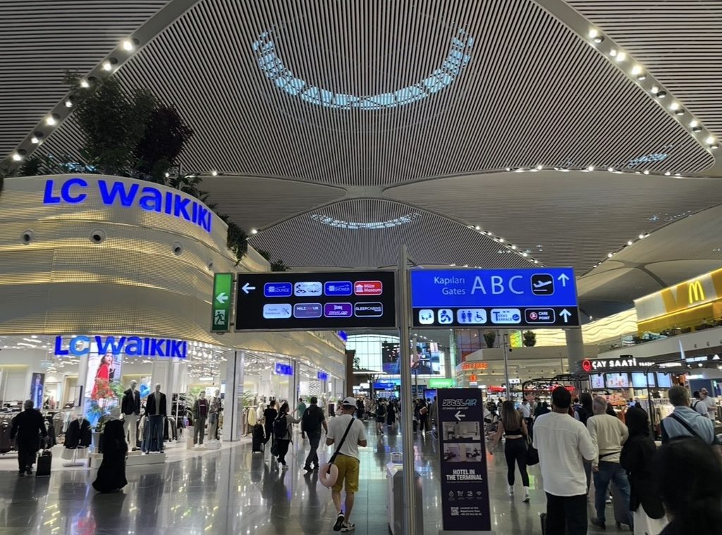 イスタンブール国際空港の広々としたターミナル内。高い天井と近代的な照明が並び、ゲート案内板やショップの前を多くの旅行者が行き交う様子を写した写真。