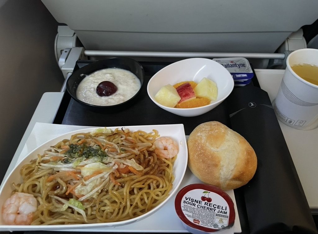 ターキッシュエアラインズの機内食として提供されたヌードル料理。エビや野菜が入った温かい麺にフルーツやデザートが添えられ、到着前の軽食として満足感のある構成になっている。