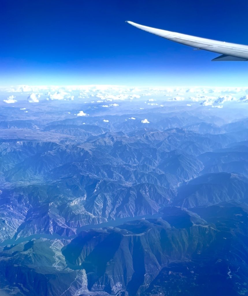 コーカサス地方の山々を上空から望む機窓風景。深い谷と険しい峰が連なり、機体の翼越しに雄大な地形が広がる。