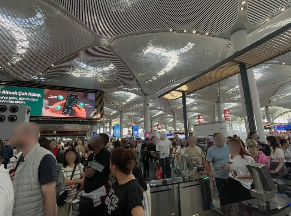 伊斯坦堡國際機場安檢門附近的擁擠景象，眾多旅客排隊等候，周圍有航班資訊螢幕與廣告，乘客依序等待辦理手續。