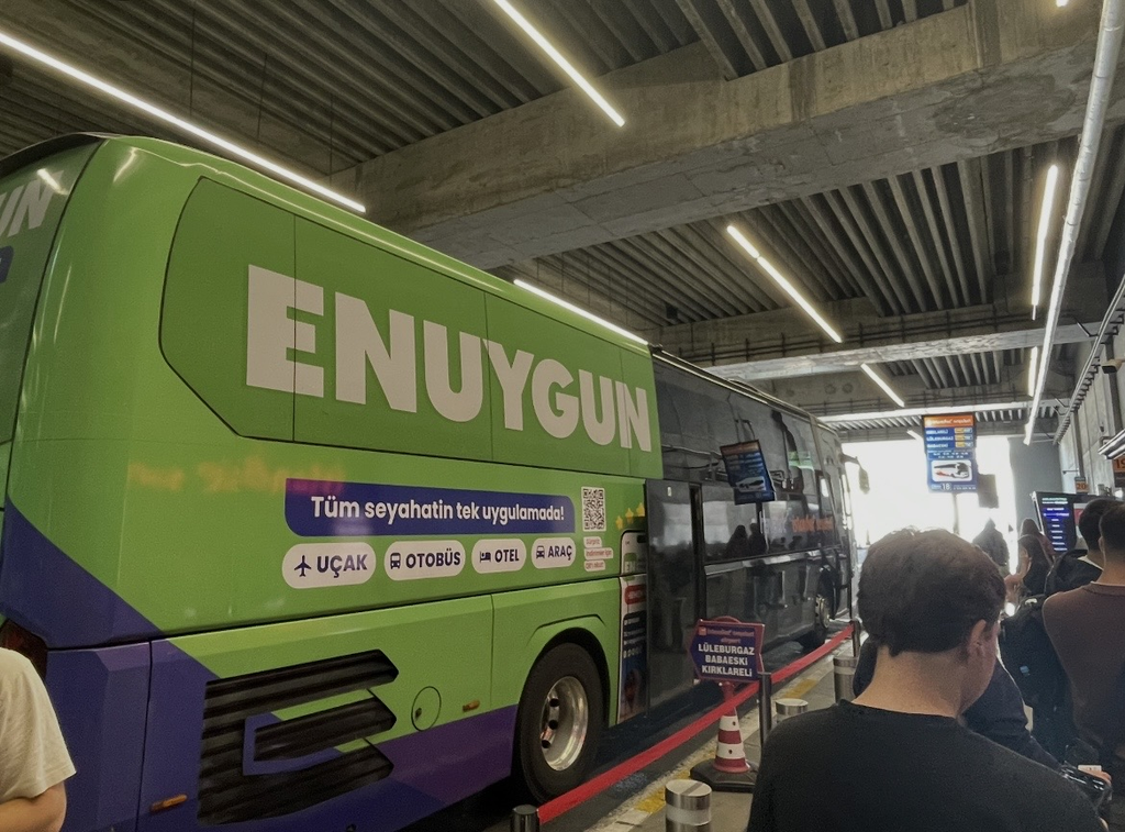 A large Havaist bus parked at the bus boarding area of Istanbul Airport, with travelers lining up to board as the airport shuttle prepares for departures into the city.