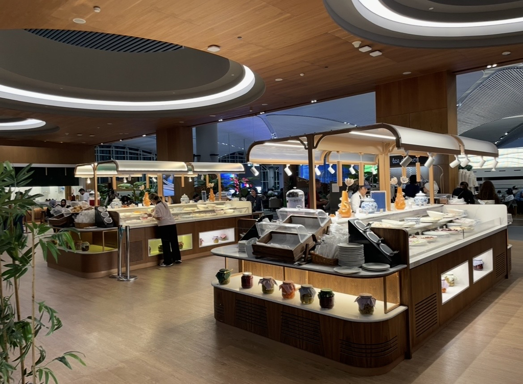 伊斯坦堡國際機場 IGA 貴賓室的自助餐區，溫暖燈光照亮的餐檯上陳列多樣料理，工作人員正在整理餐飲區的各個食物站。